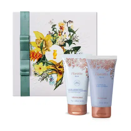 Caixa quadrada branca decorada com flores coloridas em tons de amarelo e verde e um laço verde na lateral. Em frente a ela, duas bisnagas da linha Floratta Blue, sendo um creme corporal e um gel de banho, ambos azuis com detalhes florais e tampa dourada.
