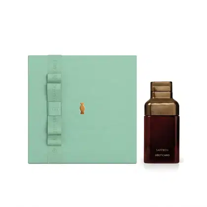 Caixa de presente verde-clara com fita no mesmo tom e o Boticário em dourado. Ao lado, vemos um frasco de vidro marrom com rótulo em letras douradas escrito "Saffron".