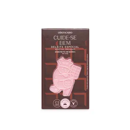 Sabonete em barra com vaca rosa estampada. Embalagem retangular marrom com texto "Cuide-se Bem Deleite Especial" e "Sabonete em Barra" em letras brancas. Selos indicando produto vegano e cruelty-free. Cores predominantes são marrom, rosa e branco.