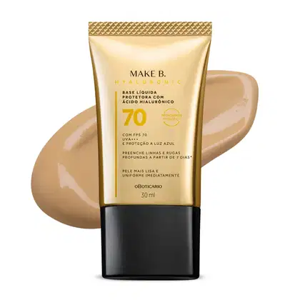 Imagem do tubo de base líquida protetora Make B. Hyaluronic Cor 20. Ele é dourado, possui tampa preta e informações em letras pretas e laranja. Atrás da embalagem está a textura do produto, mostrando sua intensidade e a real cor da base.
