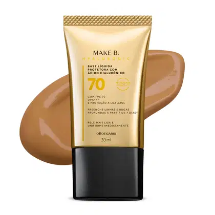 Imagem do tubo de base líquida protetora Make B. Hyaluronic Cor 60. Ele é dourado, possui tampa preta e informações em letras pretas e laranja. Atrás da embalagem está a textura do produto, mostrando sua intensidade e a real cor da base.