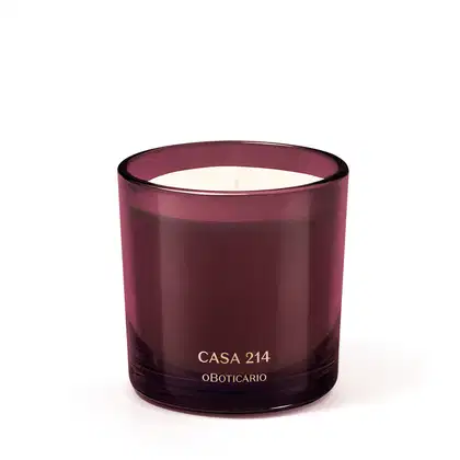 Vela aromática em recipiente de vidro de cor vinho com rótulo "Casa 214 o Boticario" em letras douradas. O recipiente possui formato cilíndrico e a vela em seu interior parece ser de cor clara.