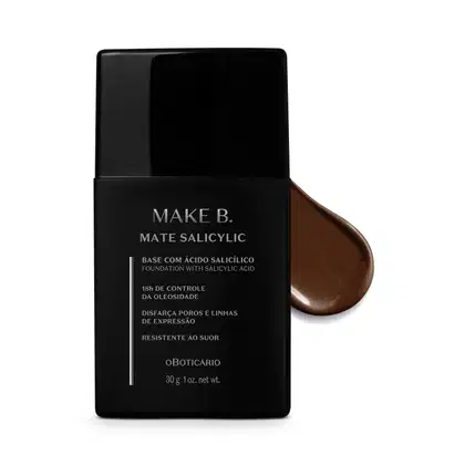 Frasco preto de base da linha Make B. Mate Salicylic com texto em branco trazendo informações do produto, como presença de ácido salicílico, controle de oleosidade e resistência ao suor. Atrás da embalagem, vemos um pouco de base marrom escuro espalhada.