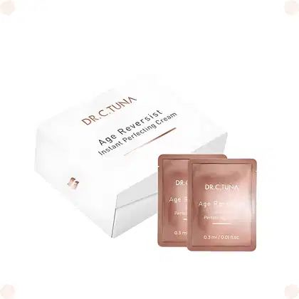 Dentro da caixa, duas embalagens marrons achatadas contendo 0,3 ml cada do creme anti-idade. As embalagens apresentam o nome da marca e do produto impresso em letras douradas.