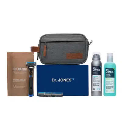A imagem mostra um kit de produtos Dr. Jones. Há um Cabo de Aparelho de Barbear, 4 lâminas de barbear, uma nécessaire cinza, um frasco de shampoo, um frasco de espuma de barbear cremosa.