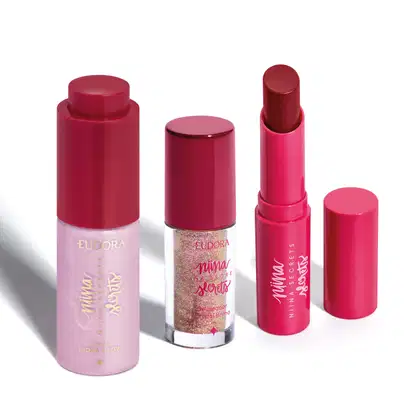 Produtos de maquiagem da linha Niina Secrets, incluindo um batom rosa, um delineador de cristal brilhante e um hidratante facial rosa. As embalagens possuem design moderno e cores predominantemente vermelhas e rosas.