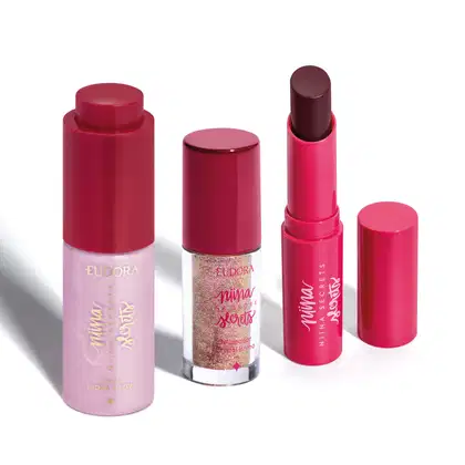 Produtos de maquiagem da linha Niina Secrets, incluindo um batom vinho, um delineador de cristal brilhante e um hidratante facial rosa. As embalagens possuem design moderno e cores predominantemente vermelhas e rosas.