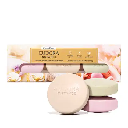 Conjunto de três sabonetes vegetais em barra perfumados. As barras têm formatos ovais e cores diferentes: rosa claro, bege e verde claro. Ao fundo, a embalagem é uma caixa retangular com imagens de doces e flores tem no centro um rótulo retangular dourado