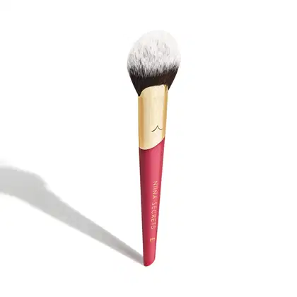A imagem mostra um pincel de maquiagem, visto de um ângulo um pouco mais elevado, sem ser exatamente de frente. Com cabo vermelho e dourado, e cerdas pretas com ponta branca. Na parte vermelha, logo da marca e linha do produto escrito em dourado.
