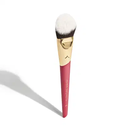 A imagem mostra um pincel de maquiagem, visto de um ângulo um pouco mais elevado, sem ser exatamente de frente. Com cabo vermelho e dourado, e cerdas pretas com ponta branca. Na parte vermelha, logo da marca e linha do produto escrito em dourado.