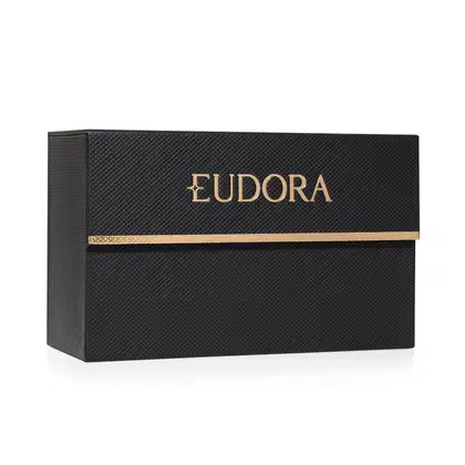 A imagem mostra o porta batom Eudora. A caixa é retangular, preta com textura rendada e o nome "Eudora" está escrito em letras douradas grandes na parte frontal com uma linha dourada horizontal abaixo.