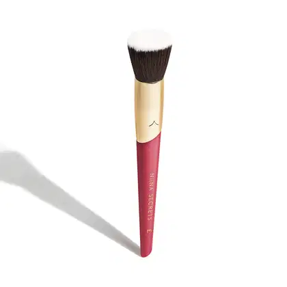 Pincel de maquiagem com cabo rosa e dourado. O cabo é cilíndrico, na cor rosa escura, com o texto "Niina Secrets" impresso. A parte superior é dourada. As cerdas do pincel são densas, corte reto e de cor marrom escura.