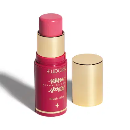 O produto apresentado é um blush stick da linha Niina Secrets da marca Eudora. Possui uma embalagem rosa com detalhes dourados, incluindo o nome da marca e da linha. O produto tem formato cilíndrico e tampa dourada.