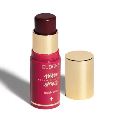 O produto apresentado é um blush em bastão da linha Niina Secrets da marca Eudora. A embalagem é composta por um tubo rosa com detalhes dourados. O produto está aberto, mostrando a cor Amora, com a tampa dourada ao lado.