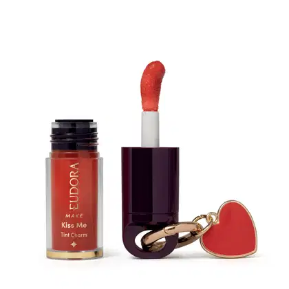 O produto de beleza Eudora Make é um batom líquido vermelho com acabamento brilhante, em embalagem cilíndrica preta e branca, acompanhado de um pingente em forma de coração vermelho.