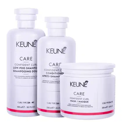 Três frascos e um pote de produtos de cuidados capilares da marca Keune, com rótulos indicando "Care Confident Curl". As embalagens são brancas com detalhes em rosa, apresentando informações sobre os tipos de cabelo e os benefícios dos produtos.