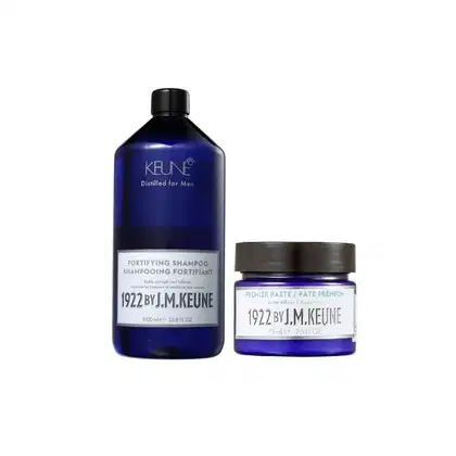 Dois produtos Keune: um frasco de shampoo fortificante e um pote de pasta premium, ambos com rótulos que indicam a marca e a linha "1922 by J.M. Keune". Apresentam embalagens de design moderno e elegante, com cores predominantes de azul e branco.