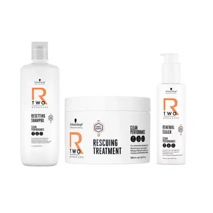 Três produtos da linha Schwarzkopf Professional R Two: frasco de shampoo, pote de tratamento e bisnaga de selador, em embalagens brancas com detalhes laranja, exibindo rótulos com o nome da linha.