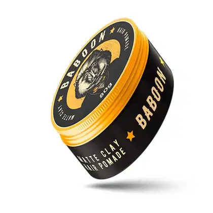 Pomada para cabelo em embalagem redonda com tampa laranja. A embalagem preta tem um rótulo circular amarelo com a ilustração de um babuíno e o texto "Baboon" em destaque. Abaixo está escrito "Mate Clay Hair Pomade" em letras menores.