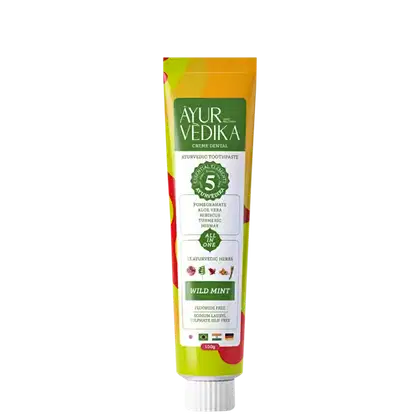 Creme dental Ayurvédico da marca AyurVedika. A embalagem é um tubo de cor amarela e verde, com rótulo destacando os ingredientes naturais como romã, aloe vera, hibisco e curcuma. O texto indica que o produto é livre de flúor e contém 25 ervas ayurvédicas.