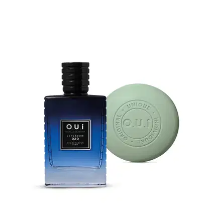 A imagem mostra dois produtos da marca O.U.i. Um deles é um frasco de eau de parfum azul escuro de formato quadrado com o nome da marca gravado na frente. O outro é um sabonete em barra de cor verde claro com a marca "o.u.i" estampada no centro.