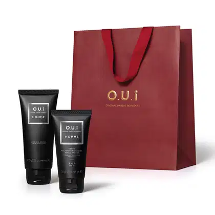 Conjunto de produtos de beleza masculina da marca O.U.I., incluindo um creme de barbear em tubo preto e um creme multifuncional em tubo cinza, acondicionados em uma sacola de compras vermelha com logotipo dourado.