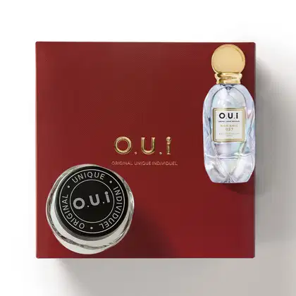 Caixa vermelha com logotipo e frasco de perfume da marca O.U.i. O frasco possui tampa dourada e rótulo com o nome da fragrância "Mon Amie". A caixa também exibe o logotipo da marca em preto e branco.
