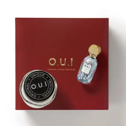 O conjunto de produtos O.U.i. inclui um frasco de perfume com tampa dourada e uma caixa vermelha com o logotipo da marca. A embalagem apresenta um design elegante e minimalista, destacando a identidade única da linha de produtos.