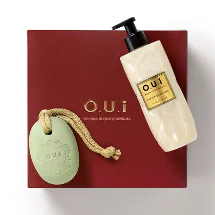 Caixa vermelha com logotipo O.U.i. e frasco de loção corporal com rótulo "Jardin de Grasse" da marca O.U.i. Embalagem e design minimalistas em tons neutros.