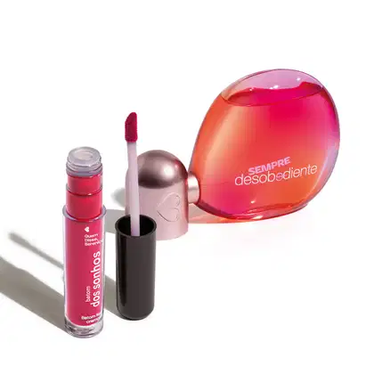 Imagem do frasco de Eau de Parfum em forma esférica, com tampa prata, líquido rosa e rótulo em letras laranja exibindo a marca "Sempre Desobediente". Ao lado, um batom líquido em tom de rosa aberto, com tampa preta, haste transparente e aplicador macio.