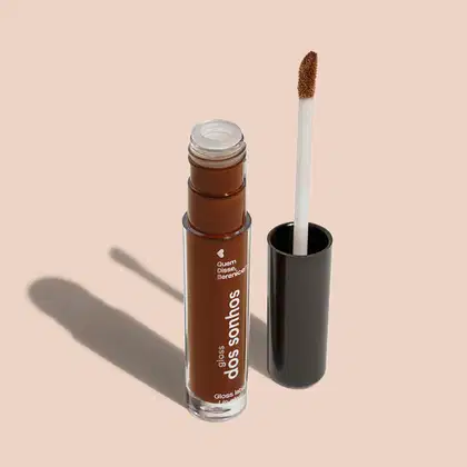 Frasco cilíndrico transparente contendo um gloss labial marrom. Rótulo na embalagem com o nome do produto "Gloss dos Sonhos" e a descrição "Quem disse, berence?". Ao lado, a tampa preta do produto, que acompanha um aplicador de espuma.