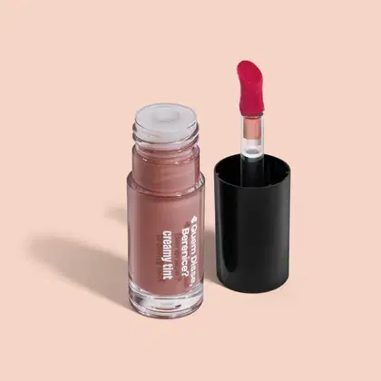 A imagem mostra um recipiente transparente de gloss labial da marca Quem Disse, Berenice? em tom de bege rosado, com aplicador vermelho, haste transparente, tampa preta, sobre superfície clara e neutra.