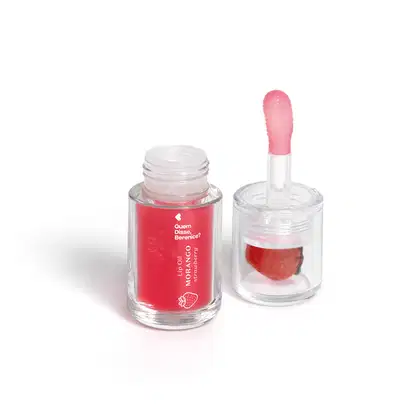 Frasco de lip oil com rótulo em vermelho e rosa, contendo um aplicador em formato de coração. Produto da marca Guem disse, Berenice, com a descrição "Lip Oil Morango" na embalagem.
