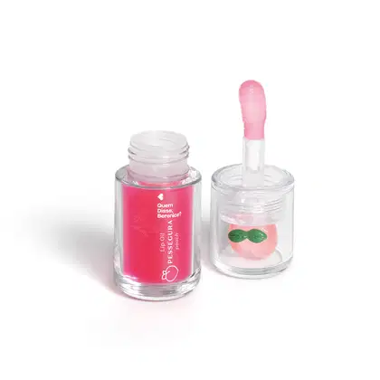 Frasco de lip oil com aplicador em forma de coração e recipiente com brilho labial em formato de pêssego, ambos em embalagens de vidro transparente com rótulos em rosa e verde.