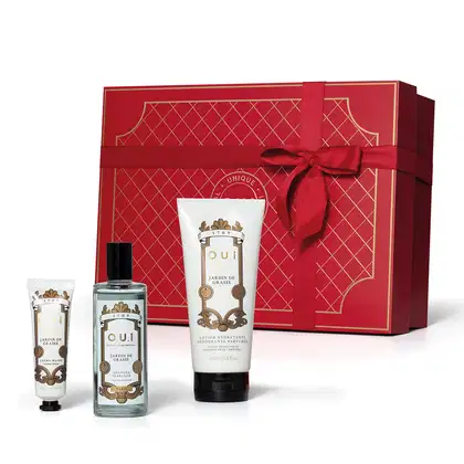 A imagem mostra um conjunto de produtos O.U.i em frente a uma bela caixa de presente vermelha. Ela traz um frasco com eau de parfum e dois tubos brancos com tampa preta, um menor com hidratante para as mãos e outro maior com hidratante para o corpo.