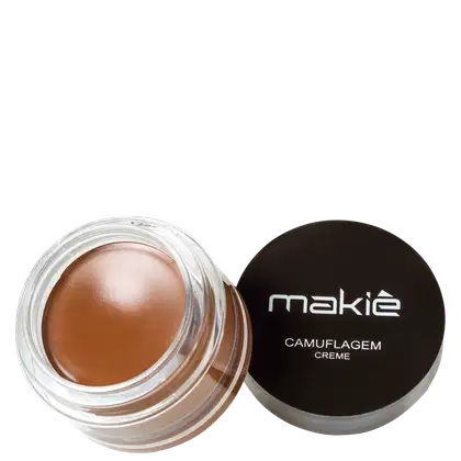 Um pote de vidro transparente contendo uma textura cremosa em tom bronzeado. A tampa preta arredondada tem a inscrição "Makiê Camuflagem Creme" em letras brancas.