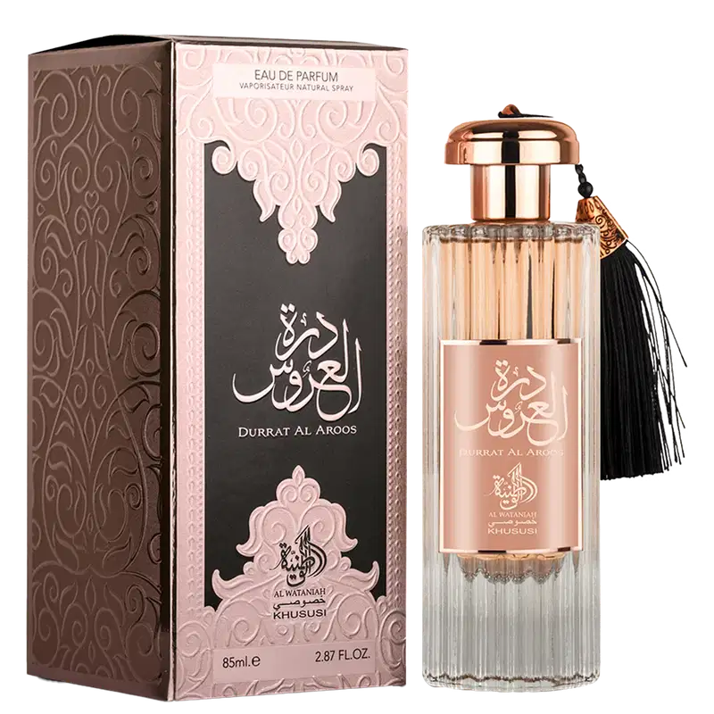 A imagem mostra o perfume e junto com a sua a embalagem, que é uma caixa retangular marrom escura com padrões ornamentais em relevo e detalhes em rosa claro. Há inscrições em árabe que parecem ser o nome do perfume "Durrat Al Aroos".