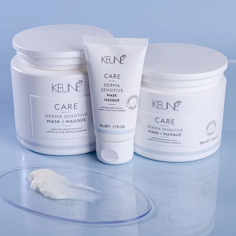 Produtos de cuidados capilares da marca Keune, incluindo máscaras de tratamento dermatologicamente sensíveis em embalagens brancas e azuis, com informações sobre os benefícios do produto impressas nos rótulos.