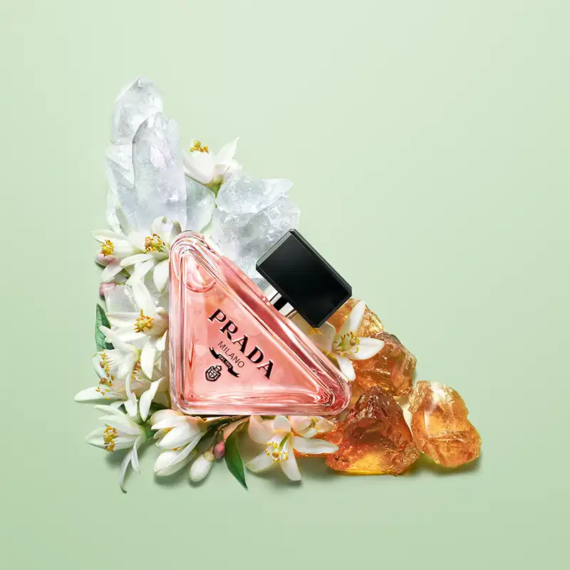 Frasco de perfume Prada com design triangular em vidro transparente, contendo líquido de cor rosa clara. O rótulo preto destaca o nome da marca "PRADA" em letras brancas.