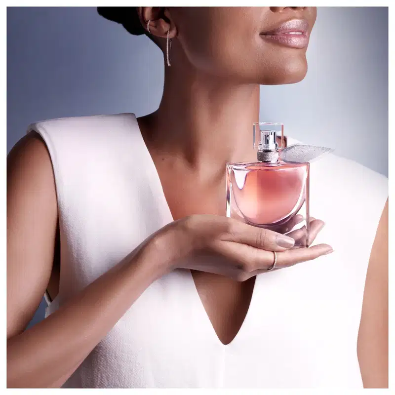 Frasco de perfume Lancôme com design elegante e tampa prateada, contendo um líquido rosado. O produto é segurado por uma mão feminina com joias discretas, destacando a sofisticação da fragrância.