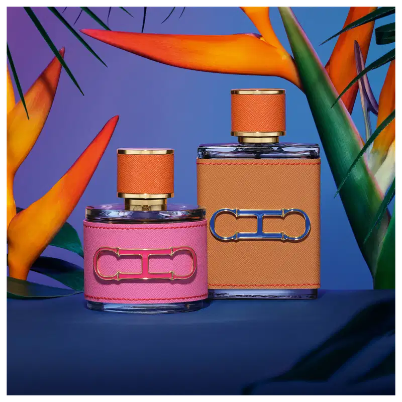 A imagem mostra dois frascos de perfumes, da mesma coleção. Um menor, arredondado, com capa rosa e tampa laranja, e um maior, retangular, com capa e tampa laranja. Ambos apresentam a logo da marca em alto relevo.