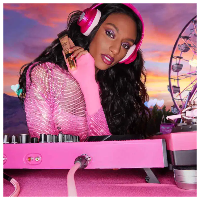 A imagem mostra uma mulher super produzida, com fone de ouvido e uma mesa de dj, ambos de cor rosa, segurando uma embalagem da base próxima ao rosto.