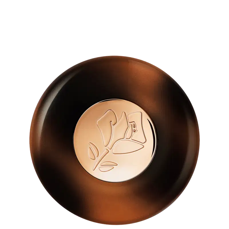 A imagem traz a embalagem do iluminador fechada em um tom marrom com detalhes em relevo na parte frontal. O frasco tem uma forma arredondada e o relevo apresenta um desenho abstrato folhado em tom mais claro. A embalagem é elegante e sofisticada.