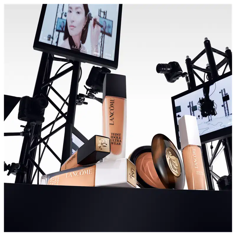 A imagem mostra alguns produtos de maquiagem Lancôme. Frascos de base líquida, na cor bege, embalagens cilíndricas com tampas pretas e brancas, com rótulos pretos e o logotipo da marca. Ao fundo alguns monitores como em um estúdio de gravação.