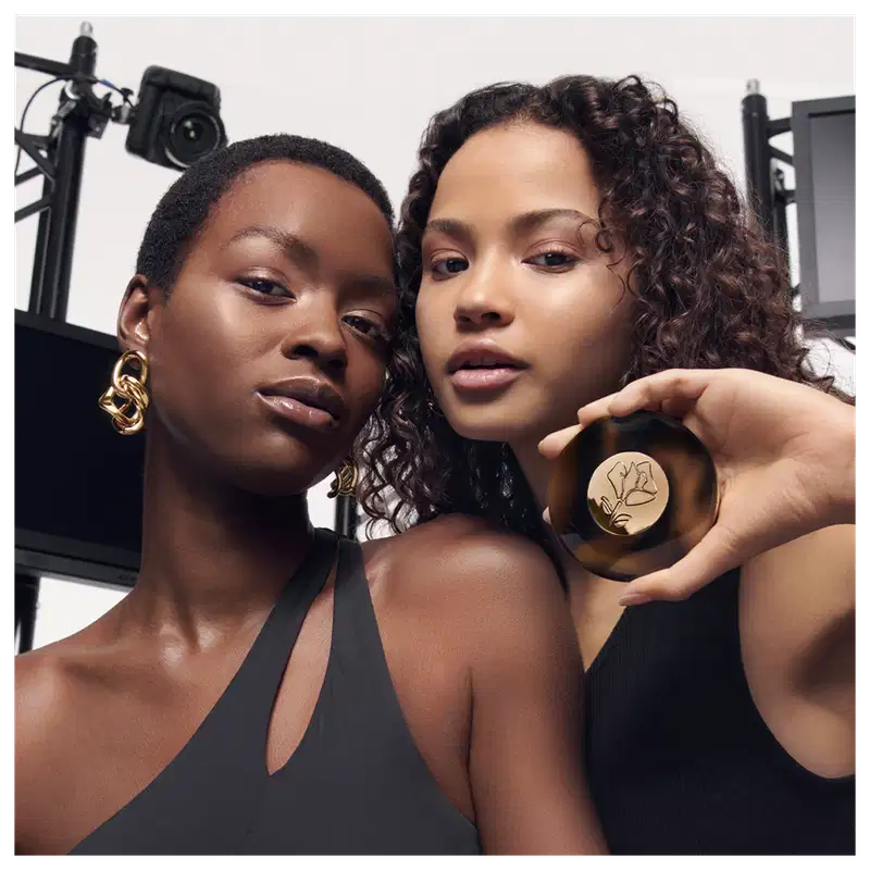 A imagem traz uma modelo de cabelo cacheado e outra de cabelo curtinho, posando em um cenário com equipamentos fotográficos ao fundo. Uma delas carregam o iluminador na mão mostrando a embalagem e a outra posa para a foto com uma expressão serena.