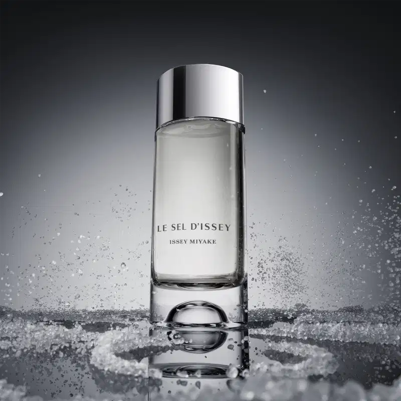 O frasco do perfume está imerso em um cenário com respingos e gotas d'água, criando um efeito visual dinâmico e sugerindo a frescura e a pureza da fragrância.