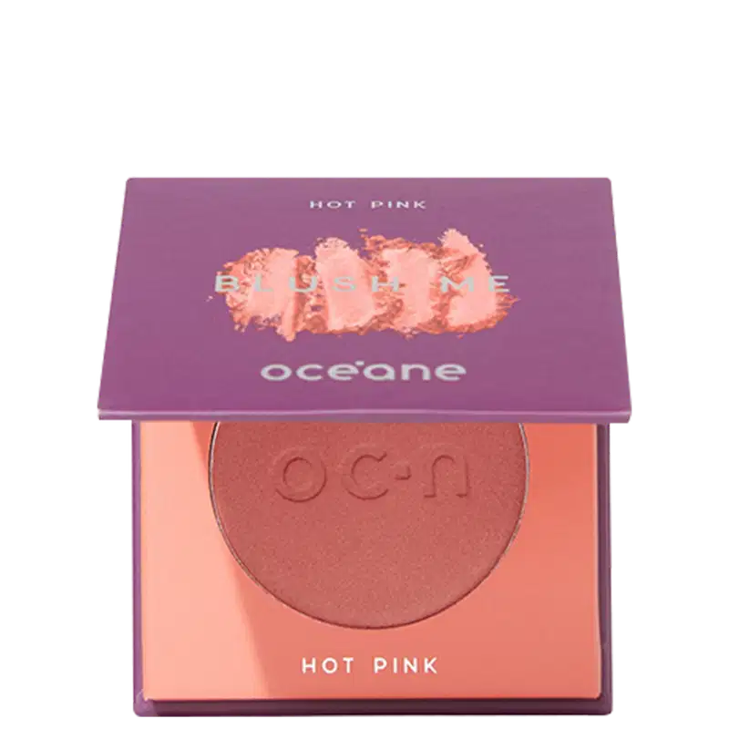 A imagem mostra o produto Blush Me da marca Océane. É uma embalagem quadrada de cor roxa que está semiaberta mostrando a cor do blush em um tom rosa com leve brilho perolado. A parte interna da embalagem é de cor pêssego claro,
