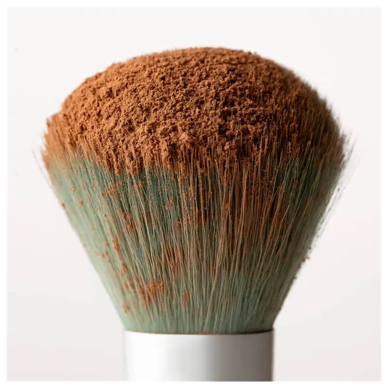Pincel de maquiagem com cerdas densas e macias de cor marrom avermelhada. O cabo é branco e a base é verde musgo com detalhes em tom ferrugem. O formato é arredondado e denso, indicando ser um pincel para aplicação de pó ou blush.