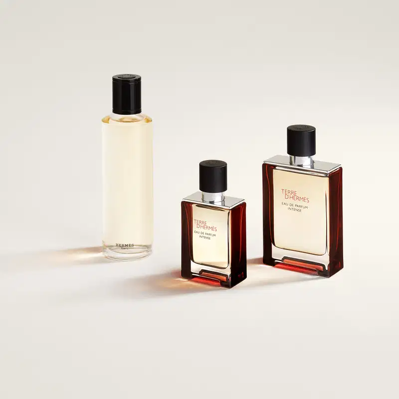 Três frascos de perfume da marca Hermès, com embalagens em tons de marrom e preto. O maior frasco contém o perfume "Terre d'Hermès Eau de Parfum Intense", enquanto os outros dois são versões menores do mesmo produto.