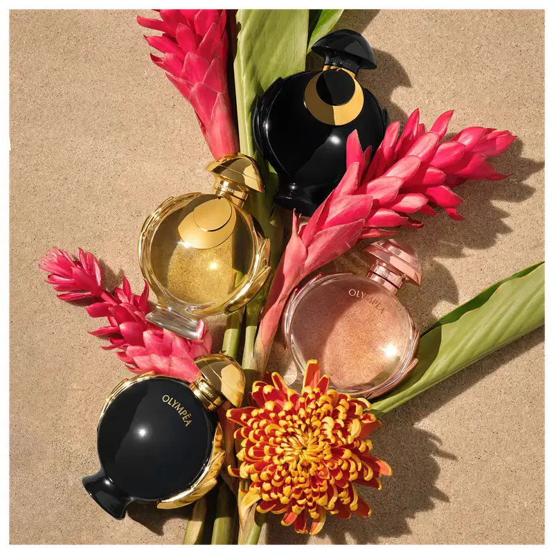 Arranjo de itens de perfumaria da marca Olympea, incluindo frascos de perfume dourados e pretos, com flores vermelhas e amarelas em um fundo bege. O cenário sugere um ambiente de beleza e luxo.